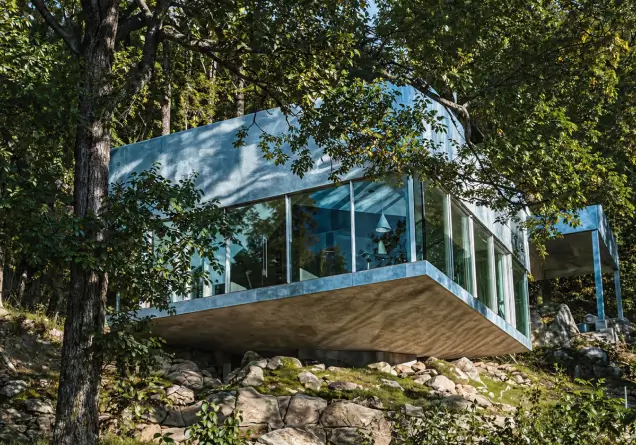 Modern glass house with concrete base elevated on rocks in a lush forested area.
