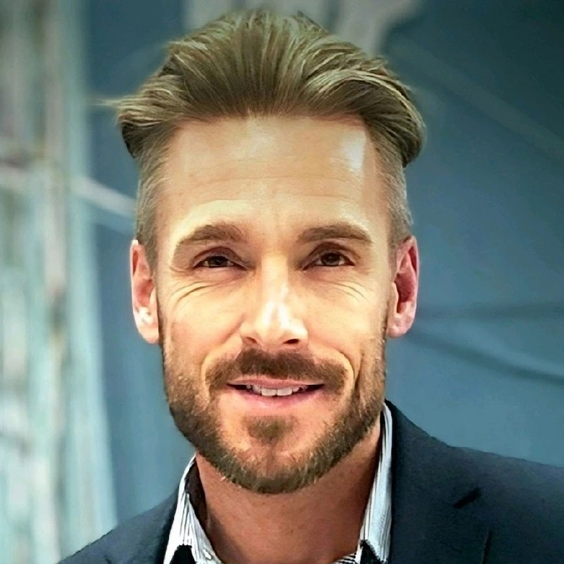 Headshot of a smiling man with light brown hair and a trimmed beard wearing a dark jacket and striped shirt.