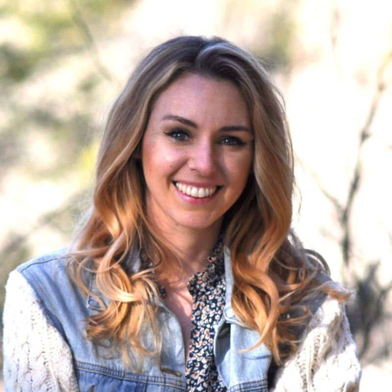 Smiling woman with wavy blonde hair wearing a denim jacket and patterned blouse outdoors.