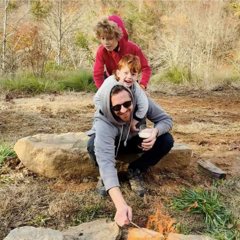 Man sitting on a rock roasting marshmallows over a small fire with two children on his back outdoors.
