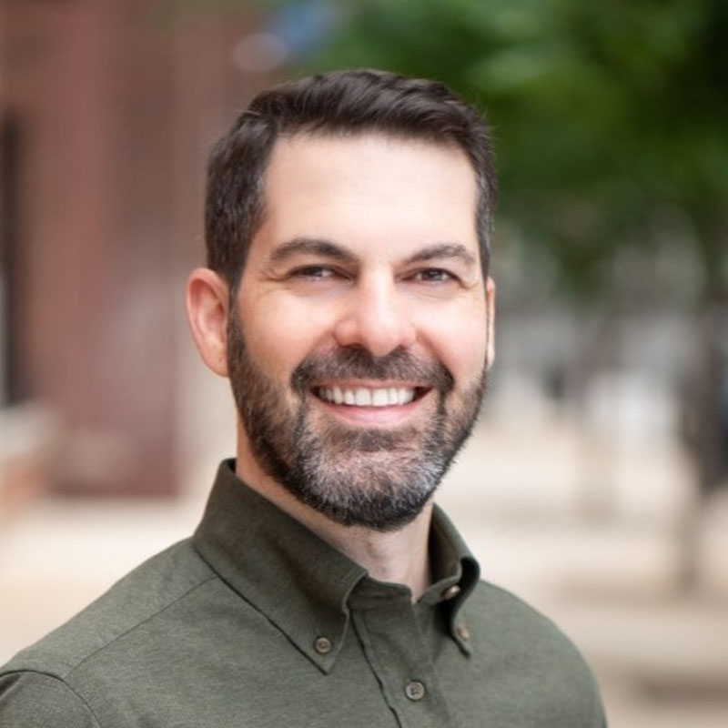 Smiling man with dark hair and beard wearing a green buttoned shirt outdoors.