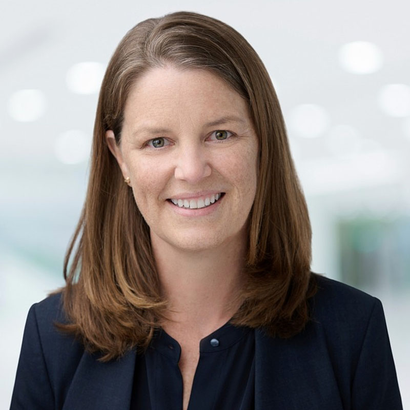 Smiling woman with shoulder-length brown hair wearing a dark blazer and blouse against a blurred background.
