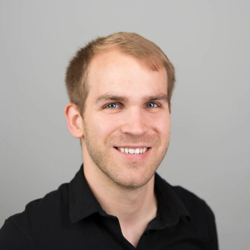 Smiling young man with short light brown hair wearing a black collared shirt against gray background.
