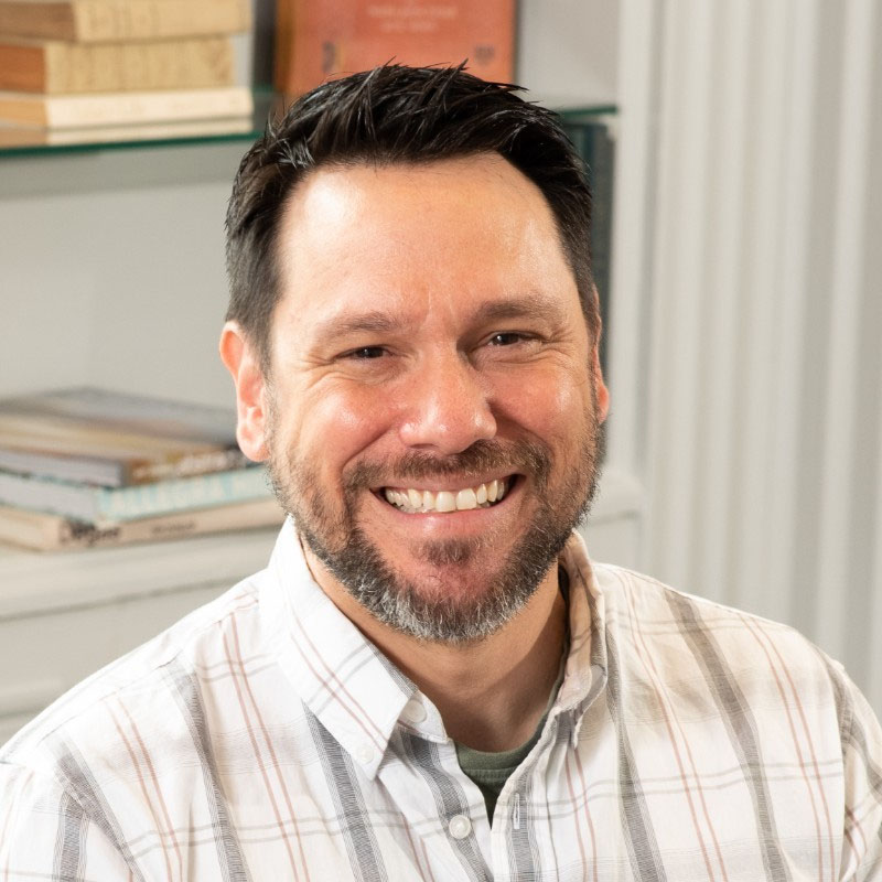Smiling man with dark hair and beard wearing a white plaid shirt in a bright room with books.