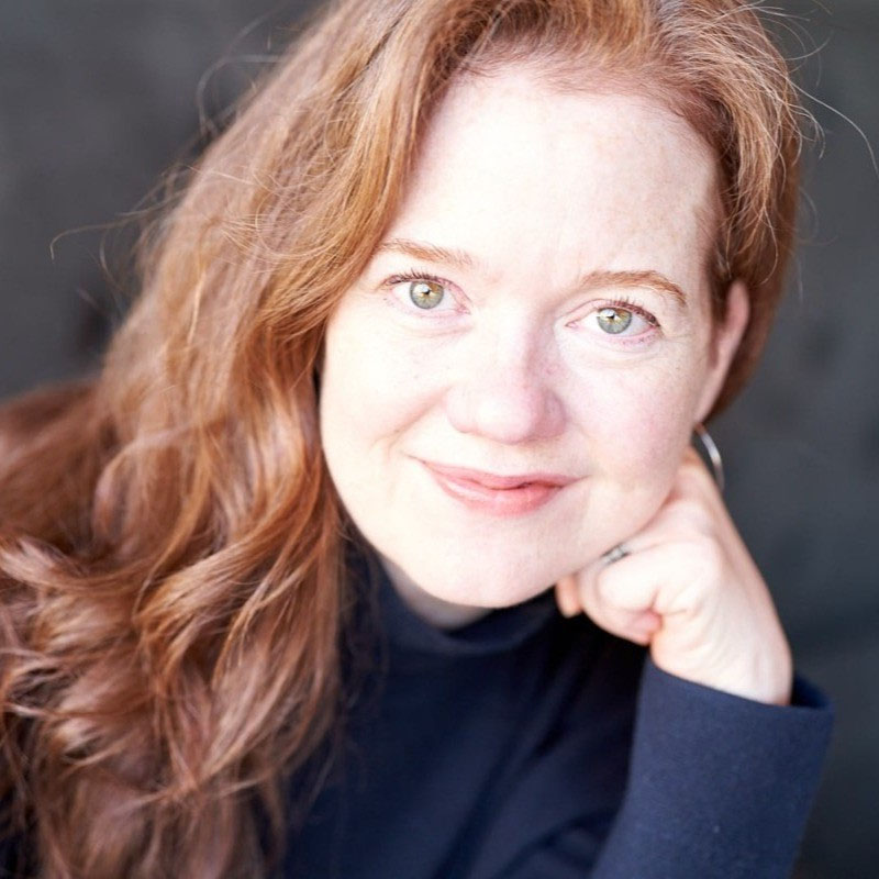 Smiling woman with long red hair resting her chin on her hand against a dark blurred background.