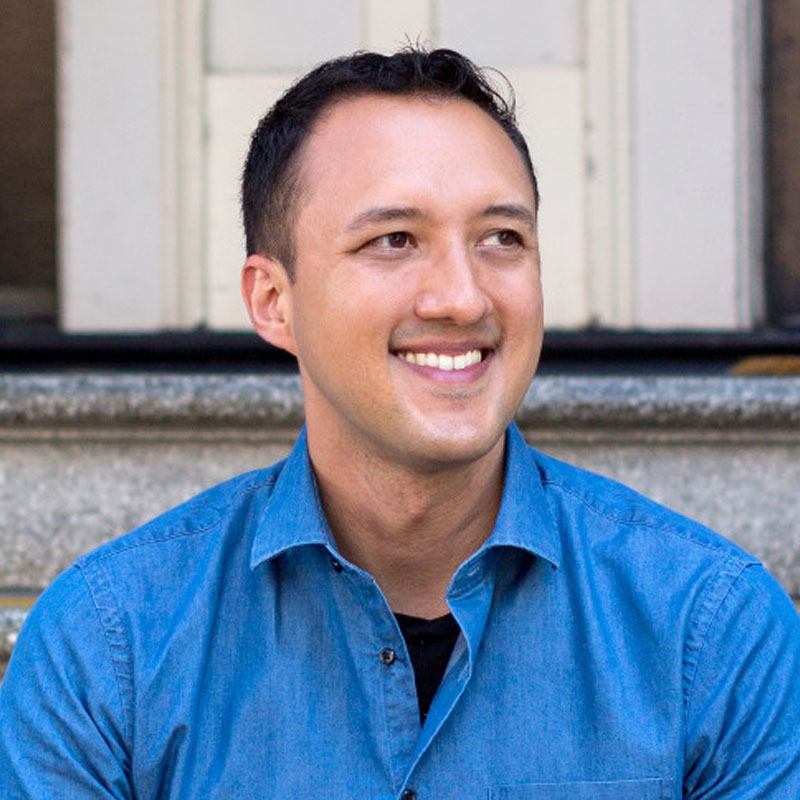 Smiling man wearing a blue button-up shirt sitting in front of a stone wall and window.