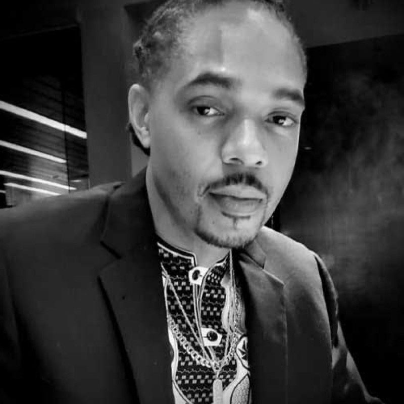 Black and white close-up portrait of a man wearing a patterned shirt and blazer indoors.