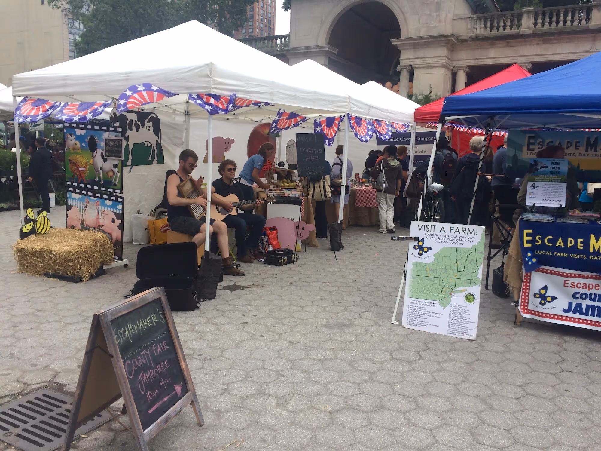 Musicians perform under one of many tents set up at an outdoor farmers market, with an a-frame sign that reads, "County Fair Jamboree 10am-4pm"