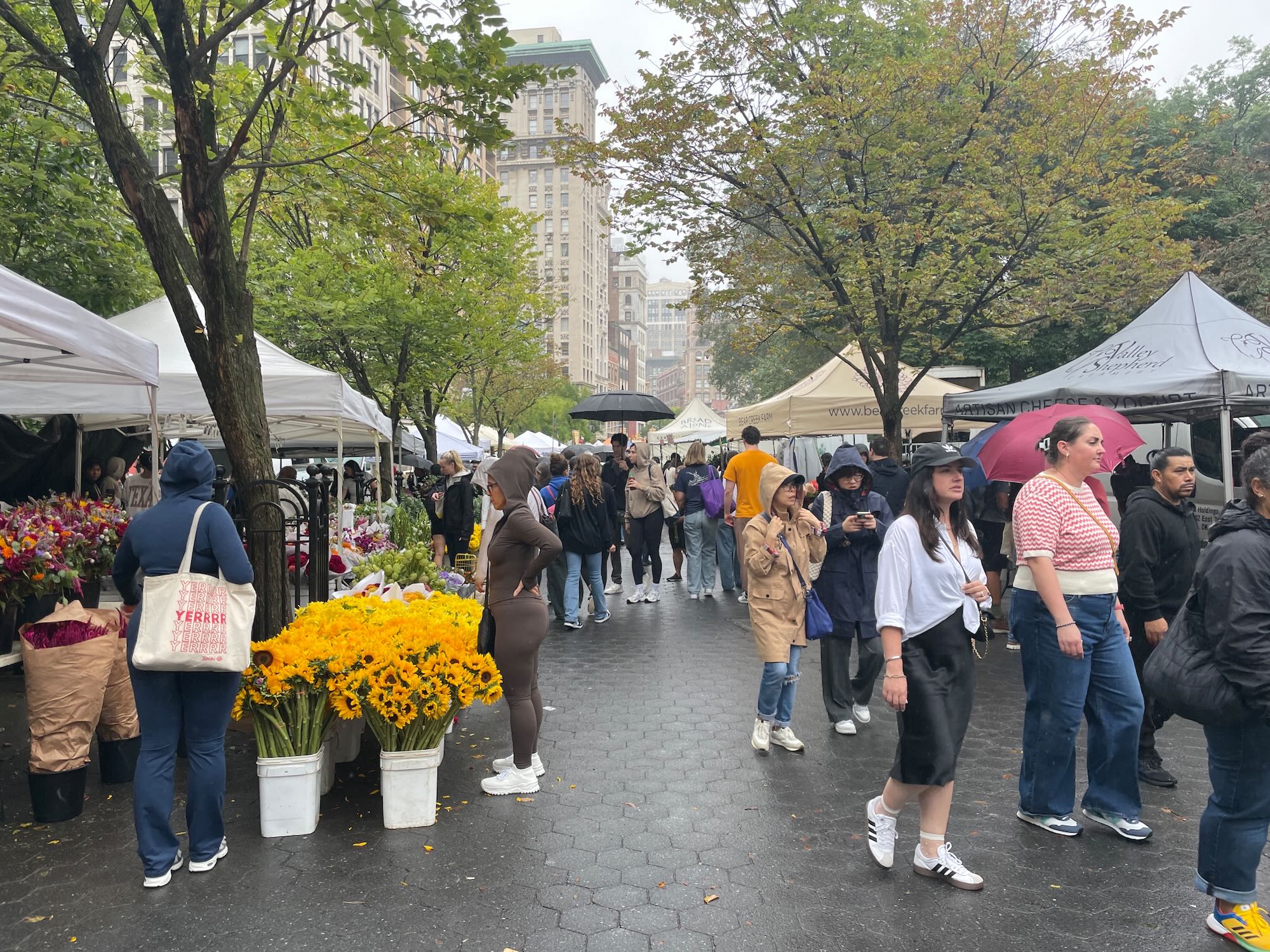 People walk through a busy outdoor farmers market, some holding umbrellas
