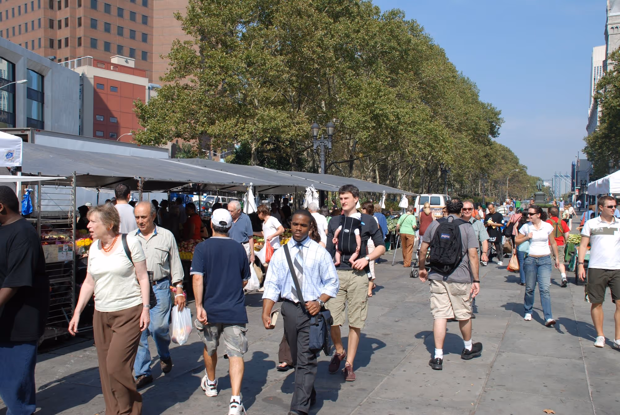 Shoppers and passersby walk through an outdoor farmers market on a sunny day