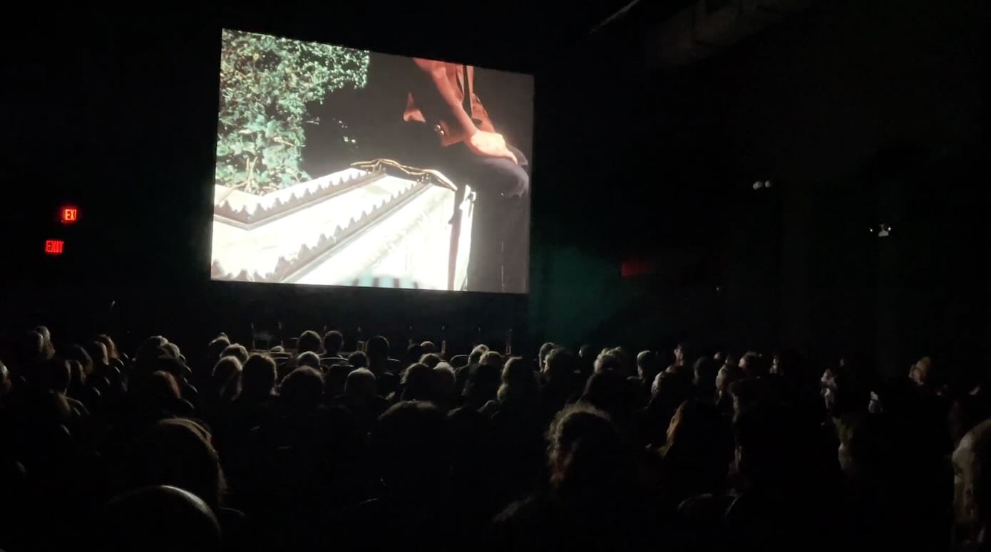 On a large movie screen in front of an audience, a man is sitting on a layer of newspaper placed on top of spikes installed on a ledge. 