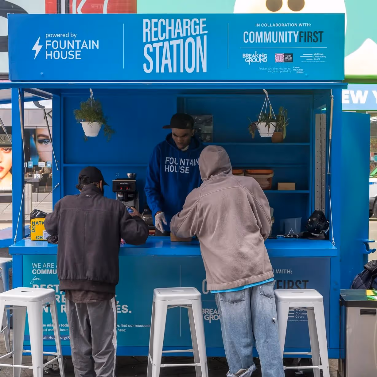 Two adults are sitting at a kiosk labeled "recharge station," in which an individual is serving coffee.