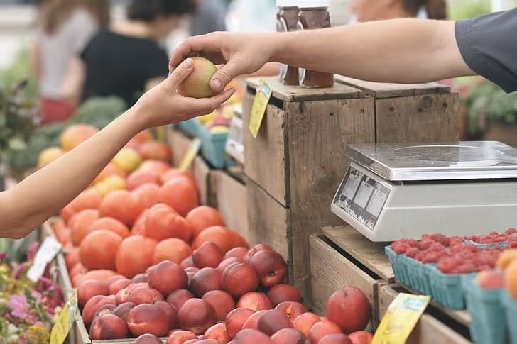 At a farmers market stand, one person's hand is placing an apple into the hand of another person.