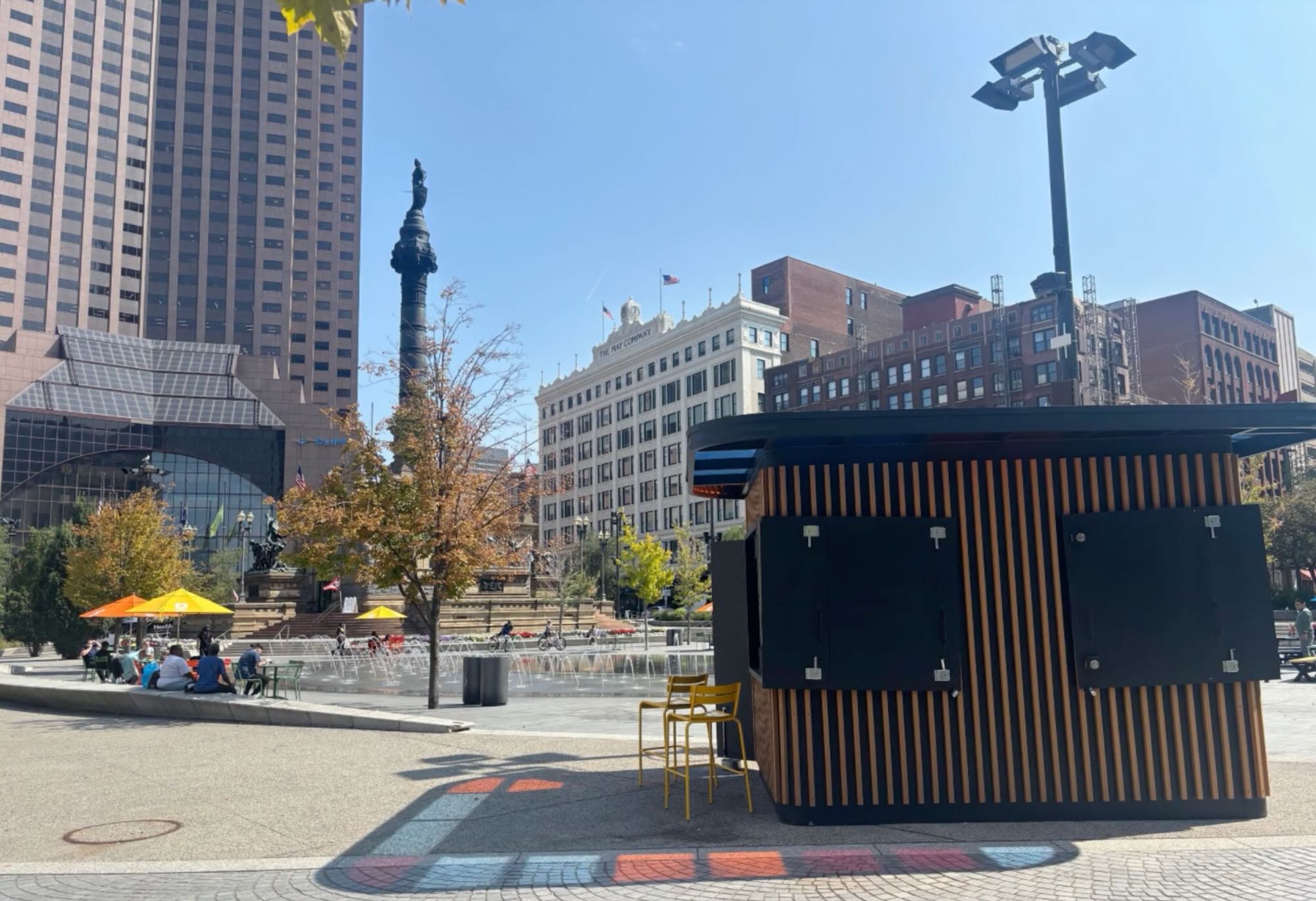 An outdoor kiosk is situated in a public square nearby a splash pad and seating area, where visitors are hanging out under the shade provided by umbrellas.