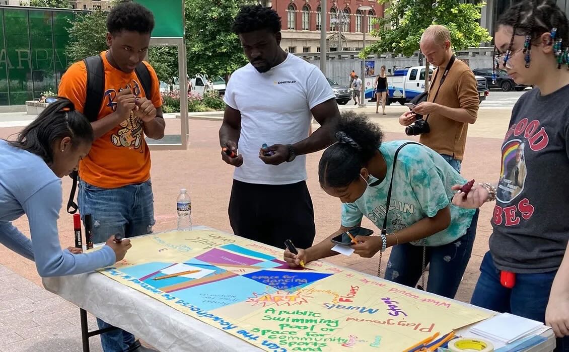 A group of individuals gather around a table outdoors, some leaning over the table to write on large pieces of paper.