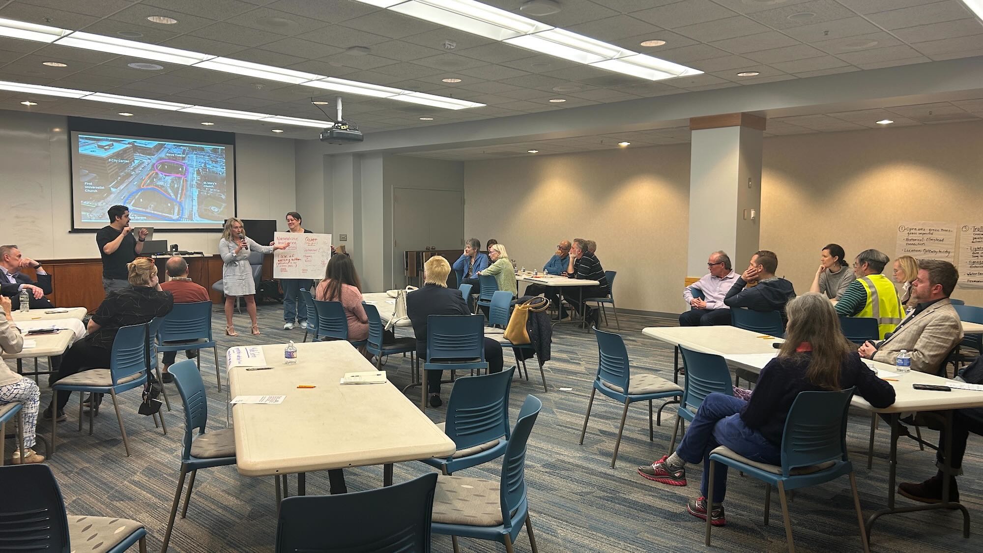 Individuals are situated in an indoor conference room, where presenters are gesturing toward a hand-written vision board.