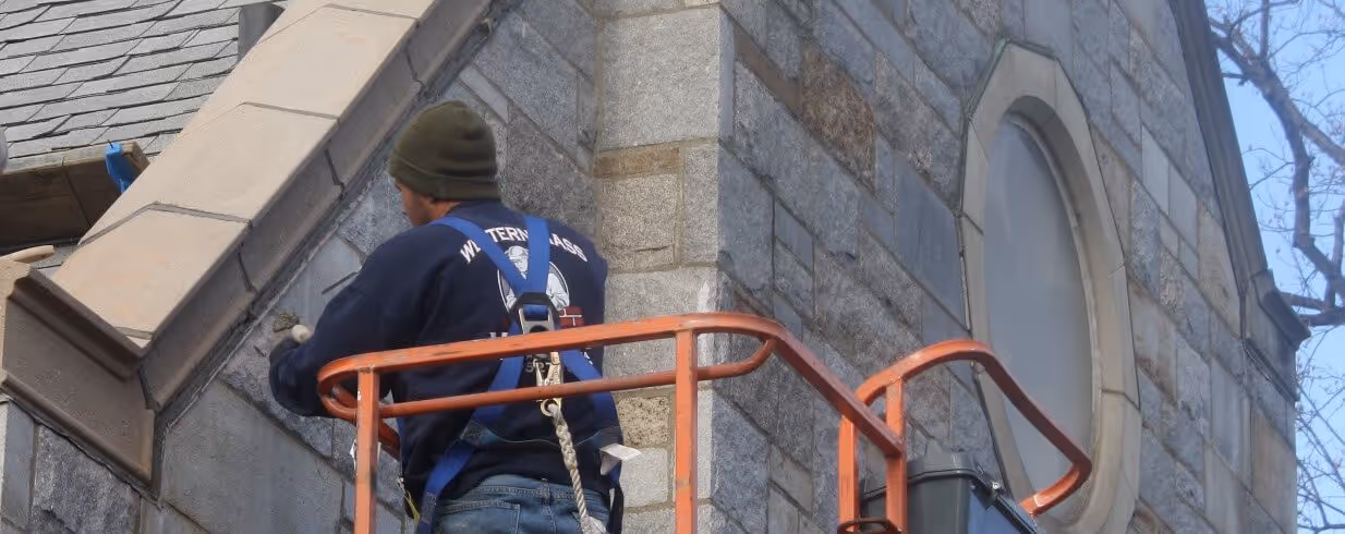 Worker wearing a harness on an orange lift platform repairing a stone building exterior near a window.