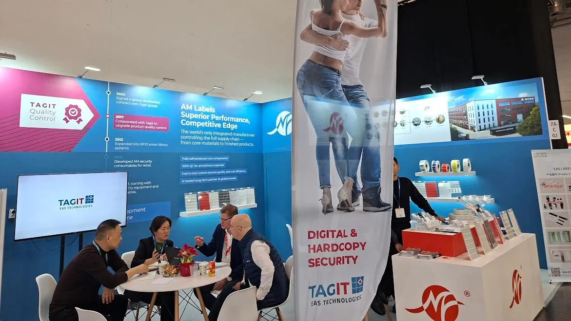 Trade show booth for TAGIT EAS Technologies featuring a meeting area with people seated around a round table, large banners promoting digital and hardcopy security and superior AM labels, and product displays on blue and white panels.