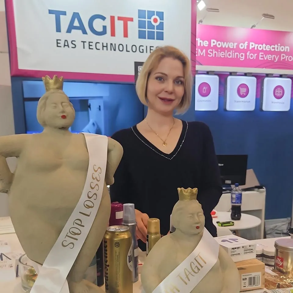 Mascha Smets standing behind a booth with two crowned statues wearing sashes that read 'STOP LOSSES' and 'TAGIT' at TAGIT EAS Technologies display.