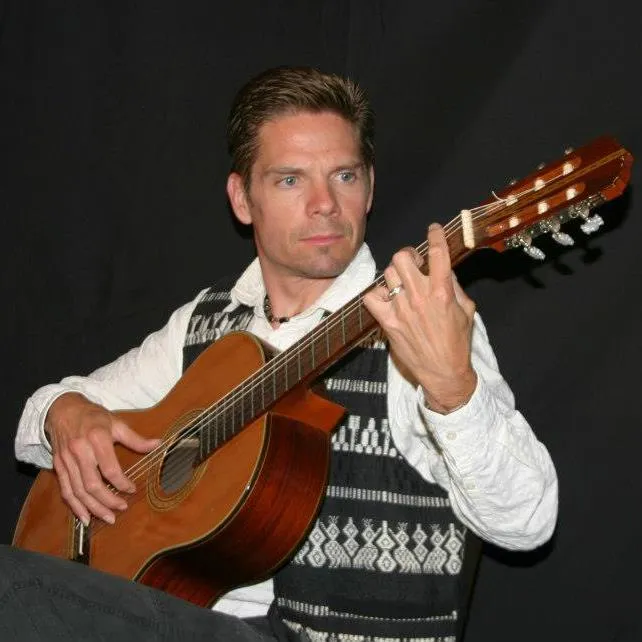 Man in white shirt and patterned vest playing a classical acoustic guitar against a dark background.