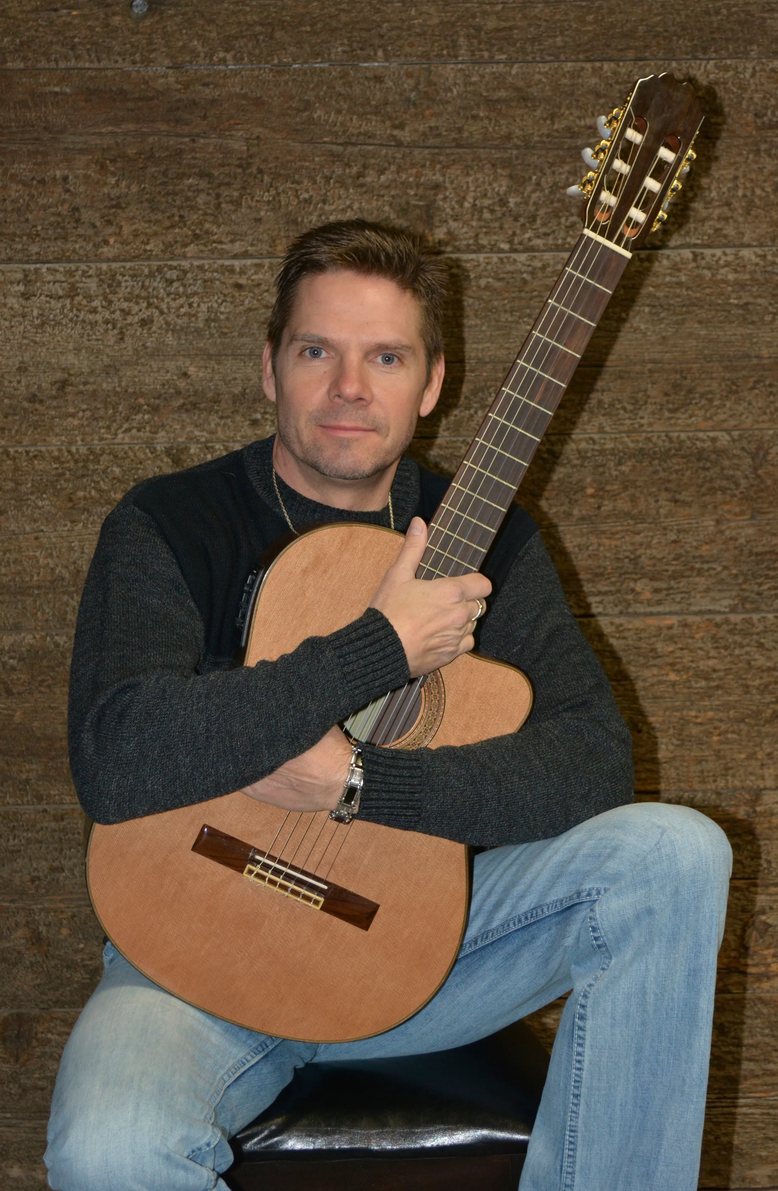 Man with short brown hair wearing a dark sweater and light blue jeans holding an acoustic guitar against a brown textured wall.