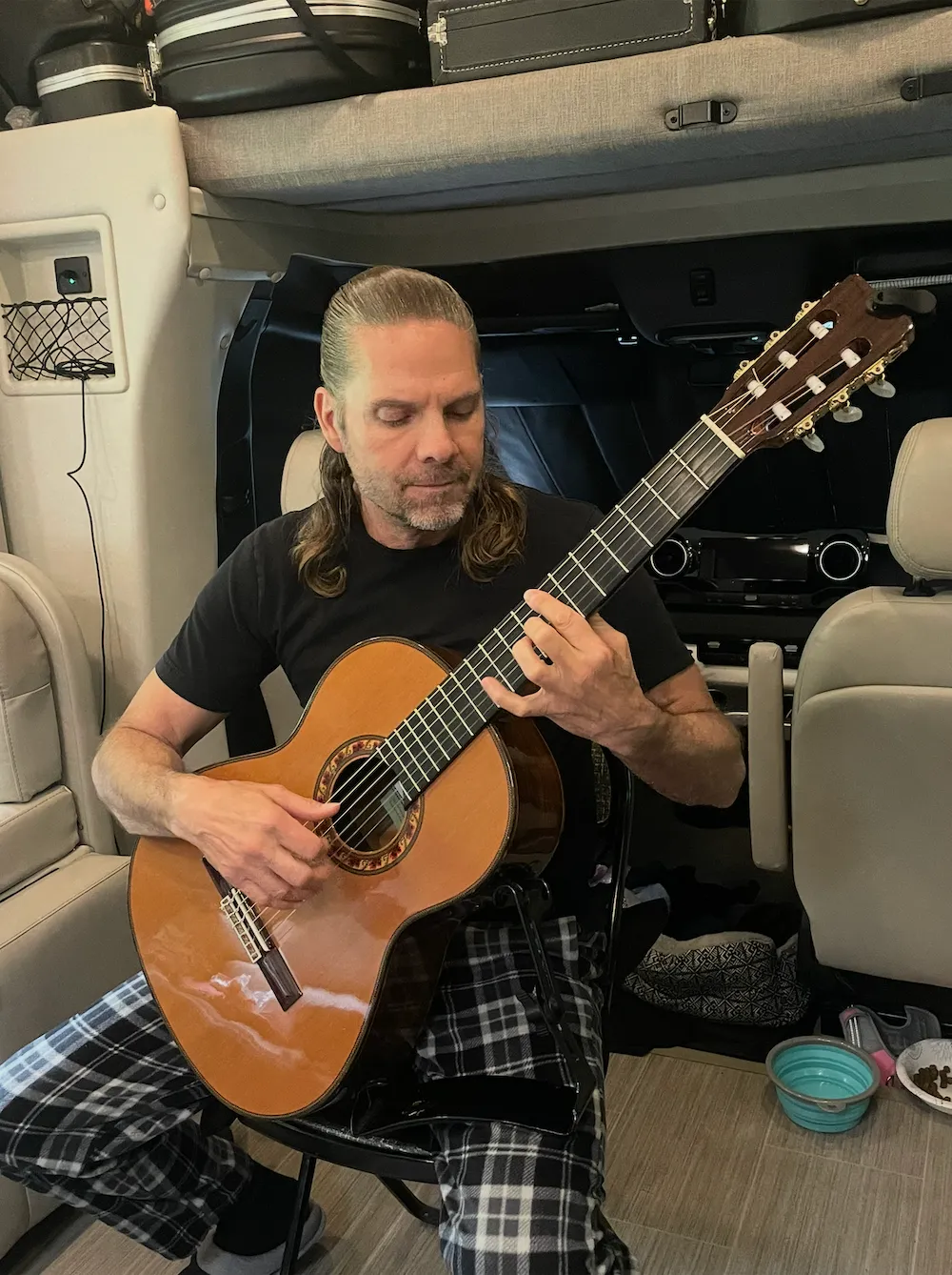 Man with long hair and plaid pants playing a classical guitar while seated inside a vehicle.