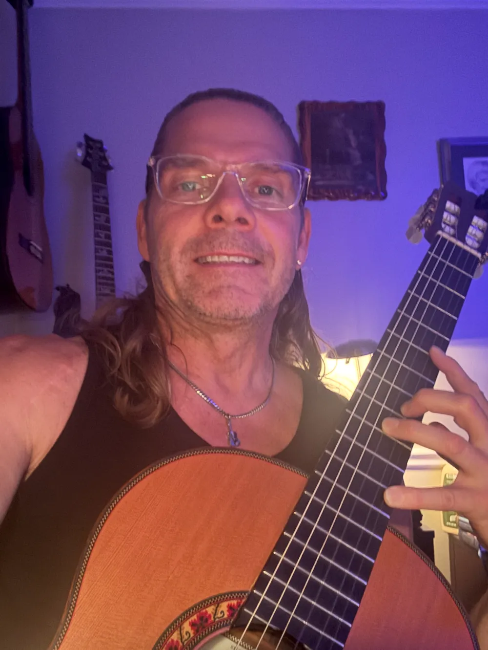 Man wearing glasses and a necklace holding a classical guitar in a room with guitars on the wall.