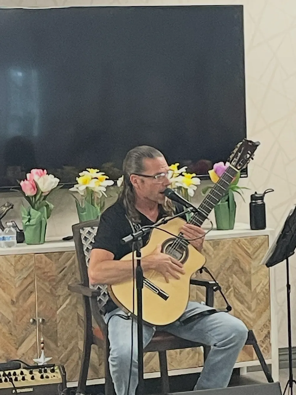 Man with long hair and glasses sitting on a chair playing an acoustic guitar and singing into a microphone indoors.