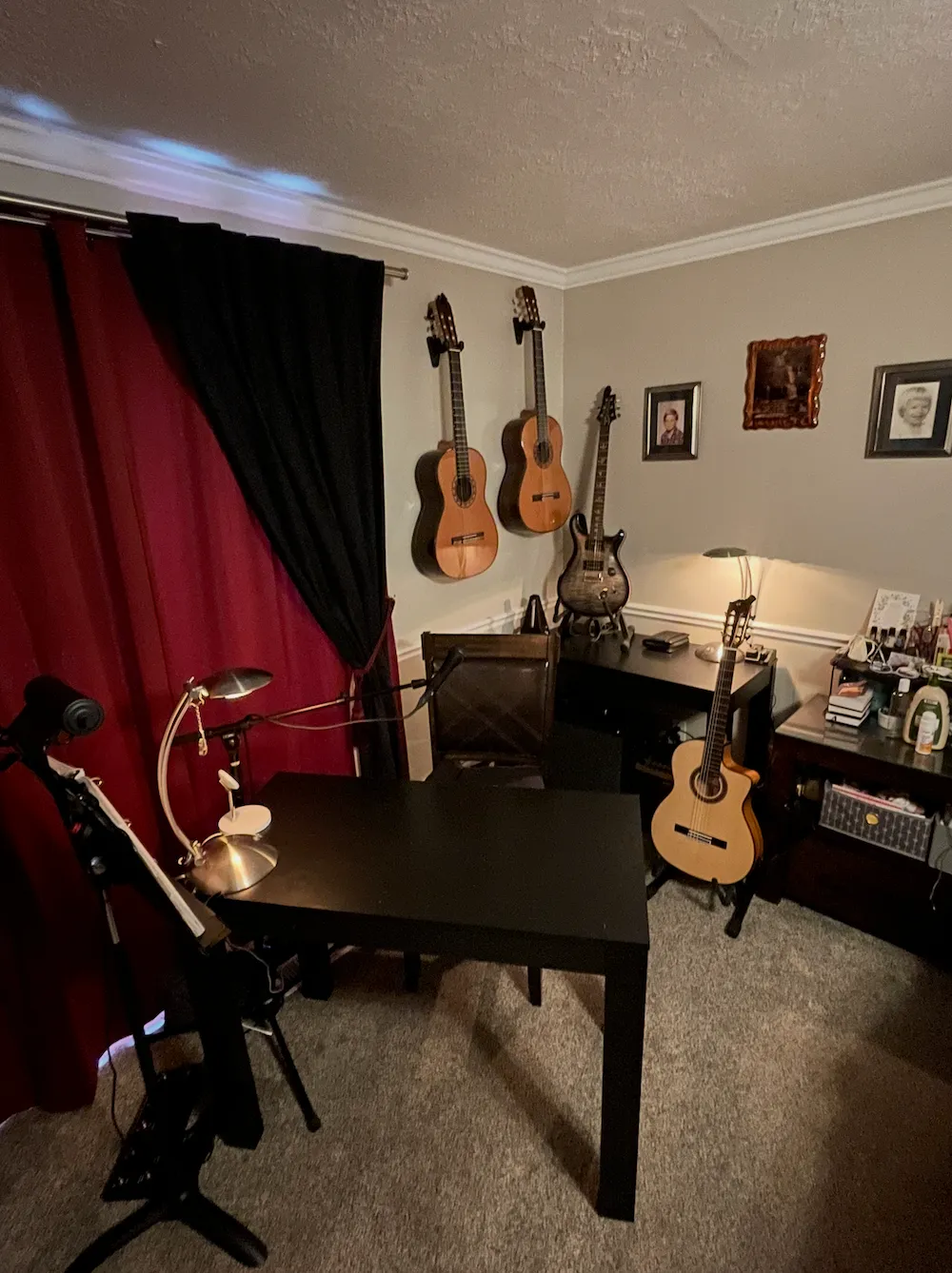 Home music room with four guitars, a black desk and chair, a desk lamp, red and black curtains, and framed photos on the wall.