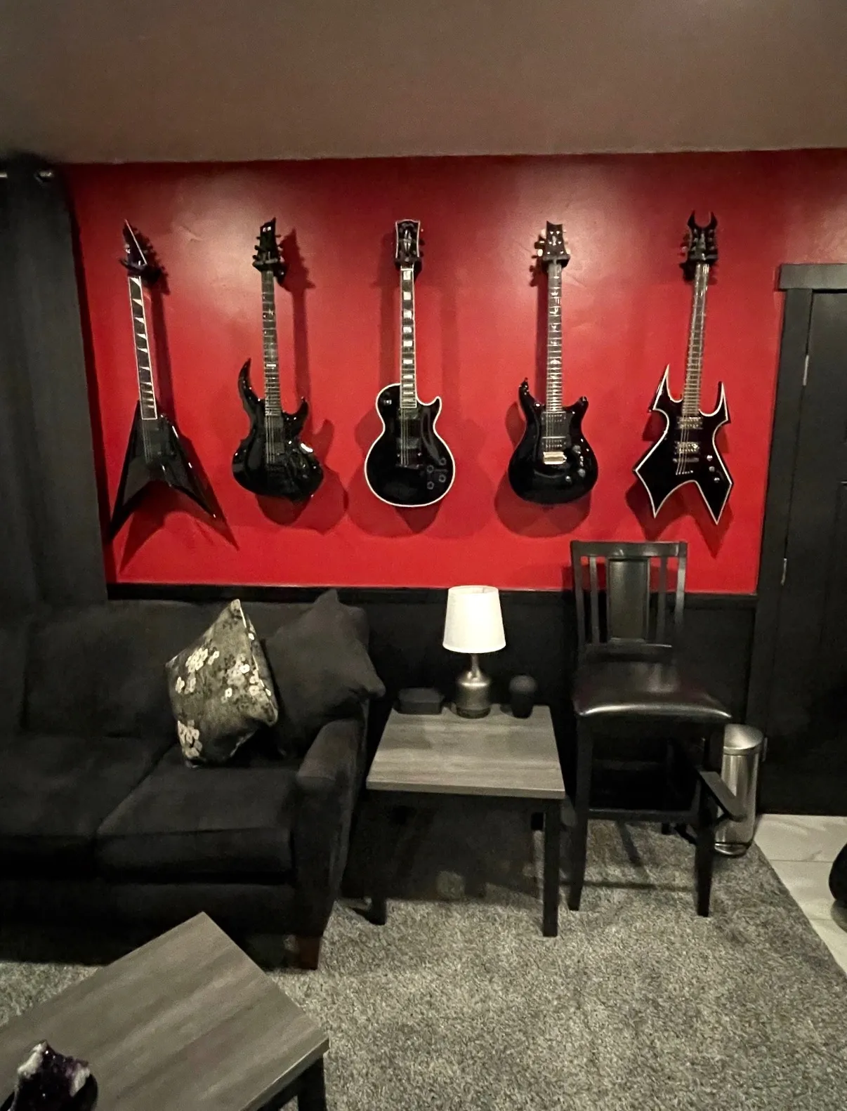 Man sitting on a camping chair playing a guitar next to a fire pit with cooking pans, a tent, and camping gear in a wooded area.