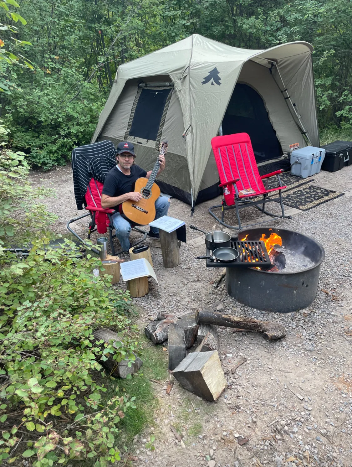Man sitting on a camping chair playing a guitar next to a fire pit with cooking pans, a tent, and camping gear in a wooded area.
