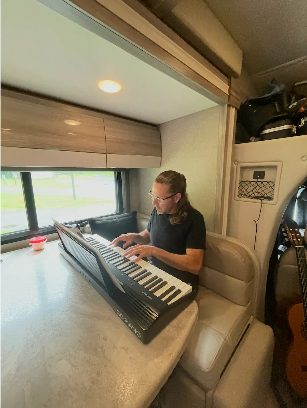 Man with glasses playing a digital piano on a table inside a camper with a guitar nearby.