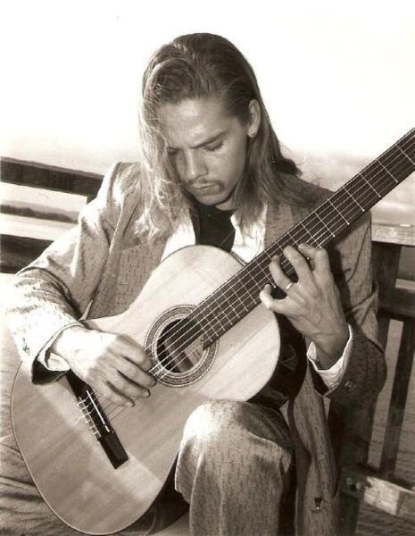 Man in white shirt and patterned vest playing a classical acoustic guitar against a dark background.