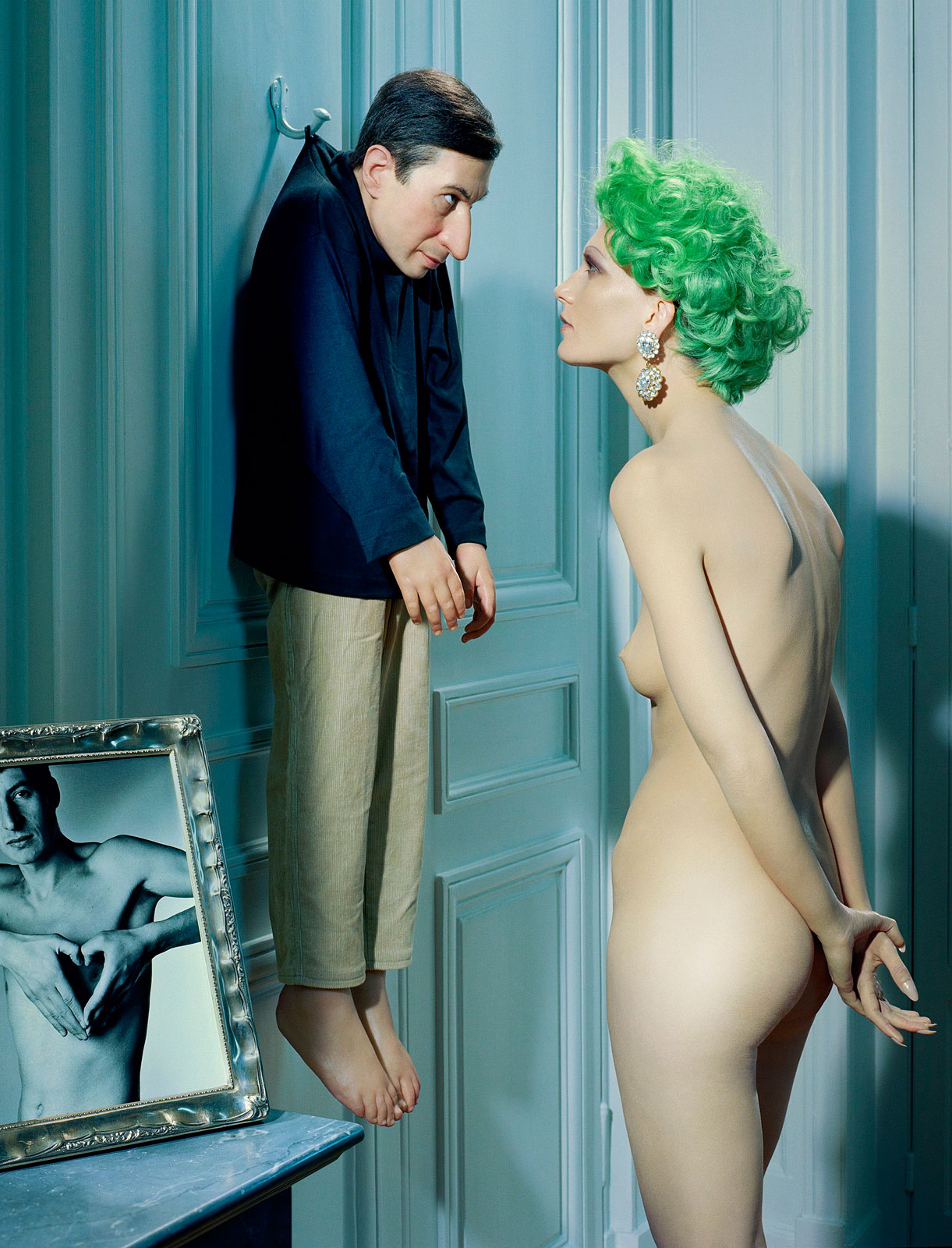 Foto van Miles Aldridge: een naakte vrouw met felgroen haar staat zijwaarts in een kamer en kijkt naar een pop of figuur in mannenkleding die aan een haak aan de muur hangt. Op de voorgrond staat een ingelijste zwart-witfoto.