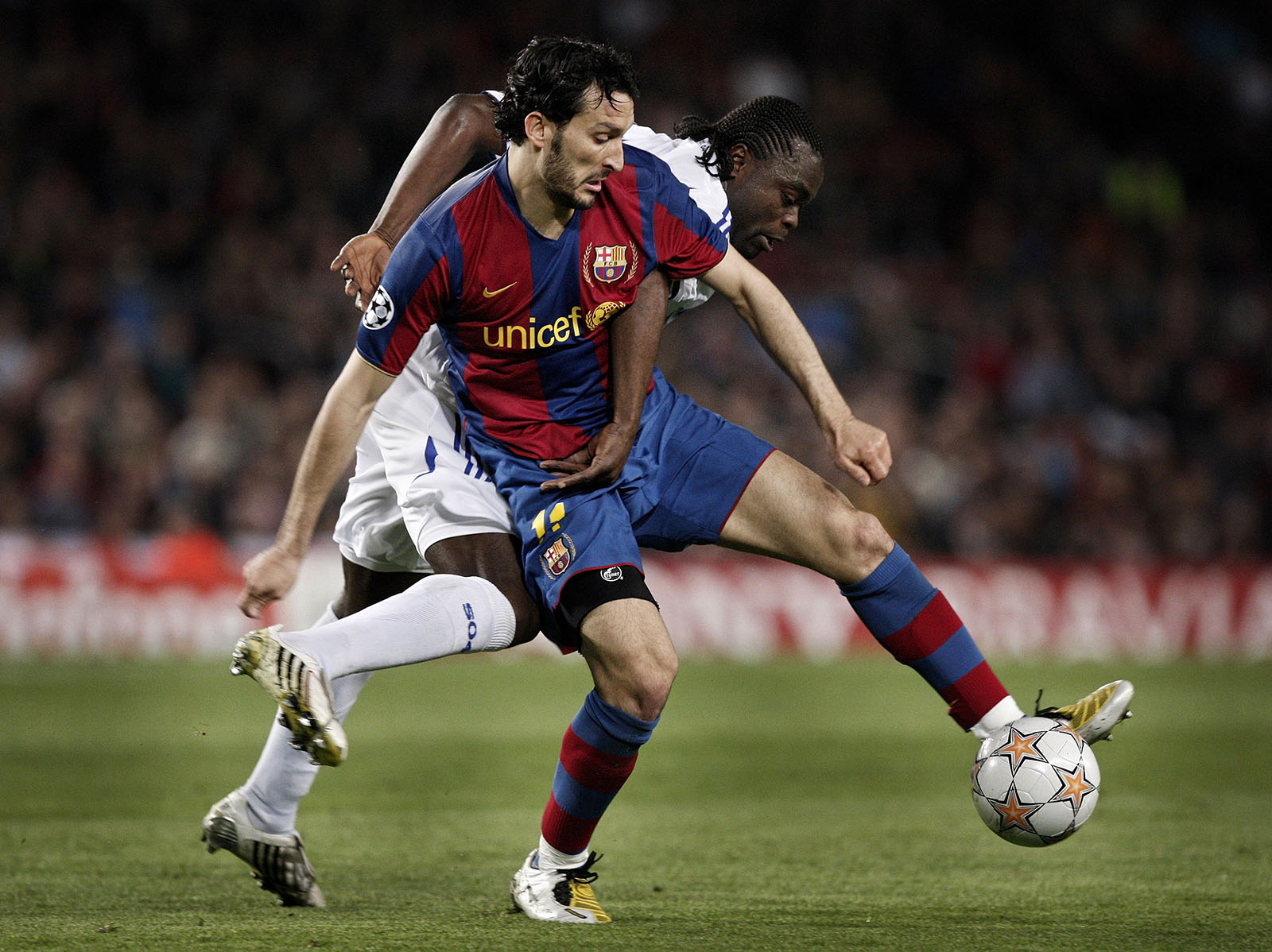 Actie tijdens een voetbalwedstrijd: speler in rood-blauw tenue schermt de bal af terwijl een tegenstander in wit-blauw verdedigt, met het veld en publiek op de achtergrond.