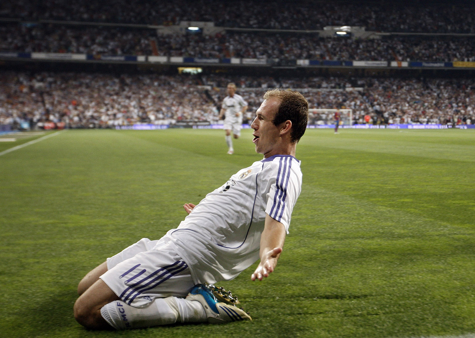 Voetballer in wit tenue glijdt juichend op zijn knieën over het veld na een doelpunt in een stadion vol toeschouwers.