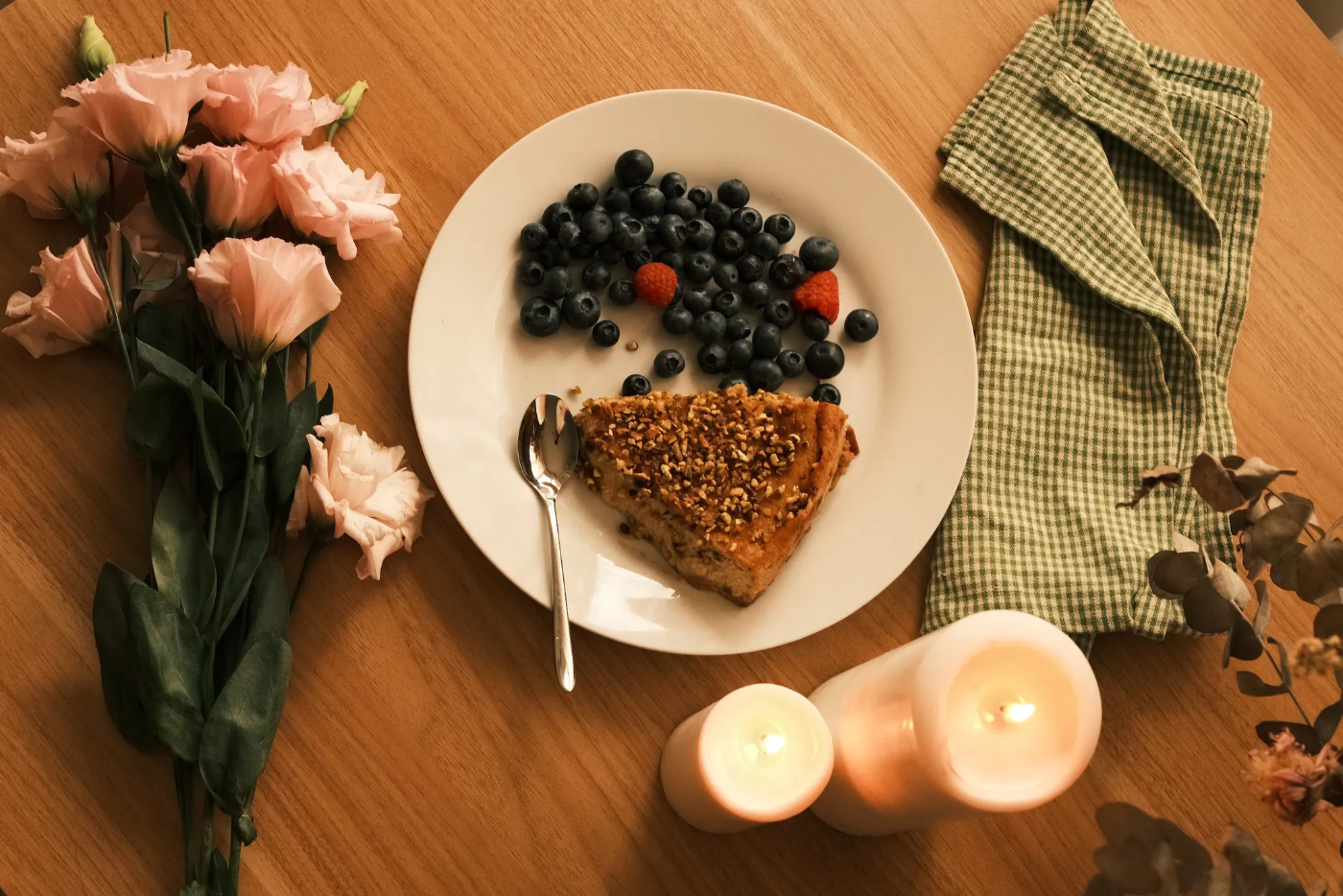 Stilleven met een stuk notentaart, blauwe bessen en frambozen op een wit bord, met kaarsen en bloemen op een houten tafel.