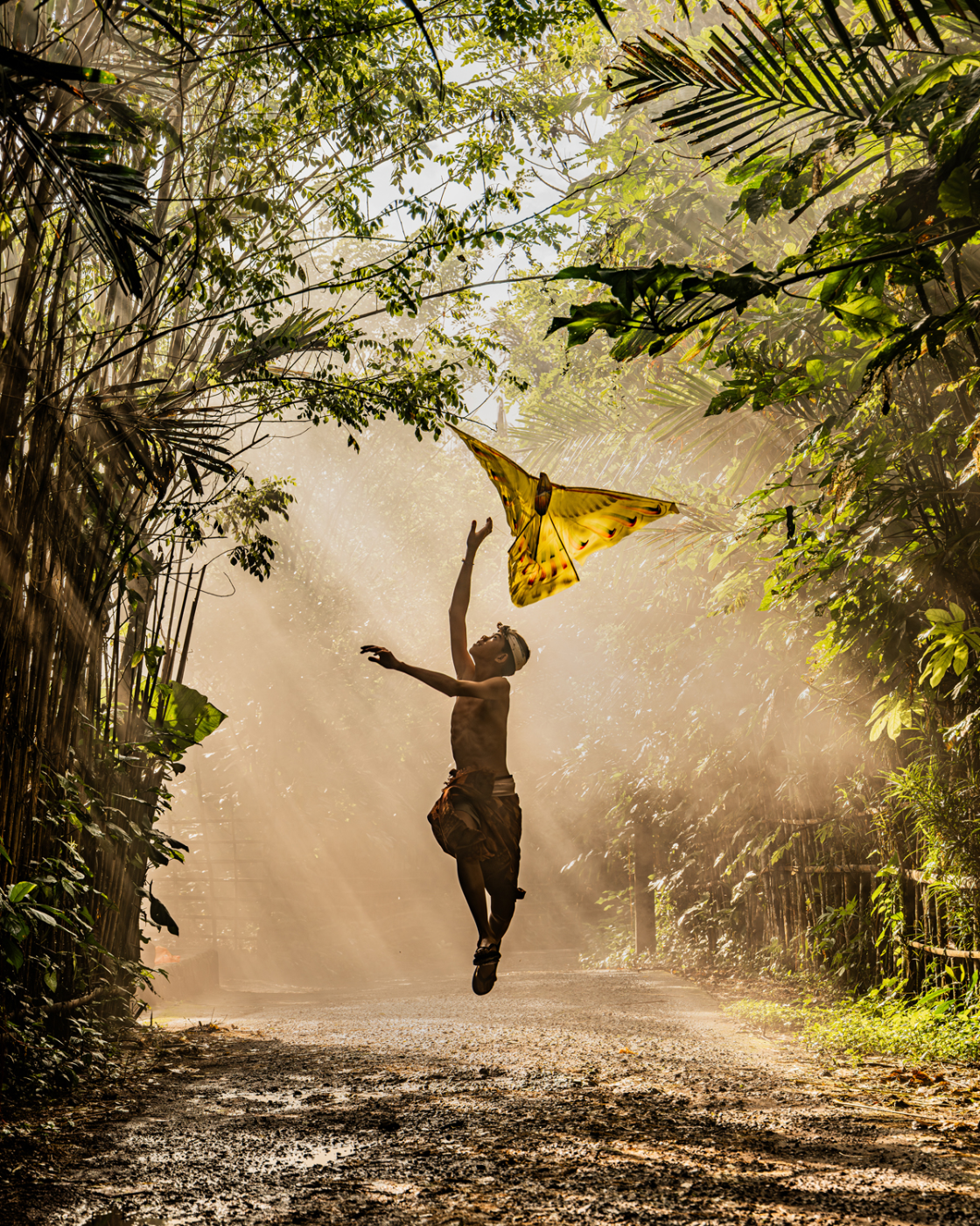 Jongen springt in het ochtendlicht om een zelfgemaakte vlieger te grijpen op een landweggetje in Bali, vastgelegd met tegenlicht en zachte achtergrondonscherpte