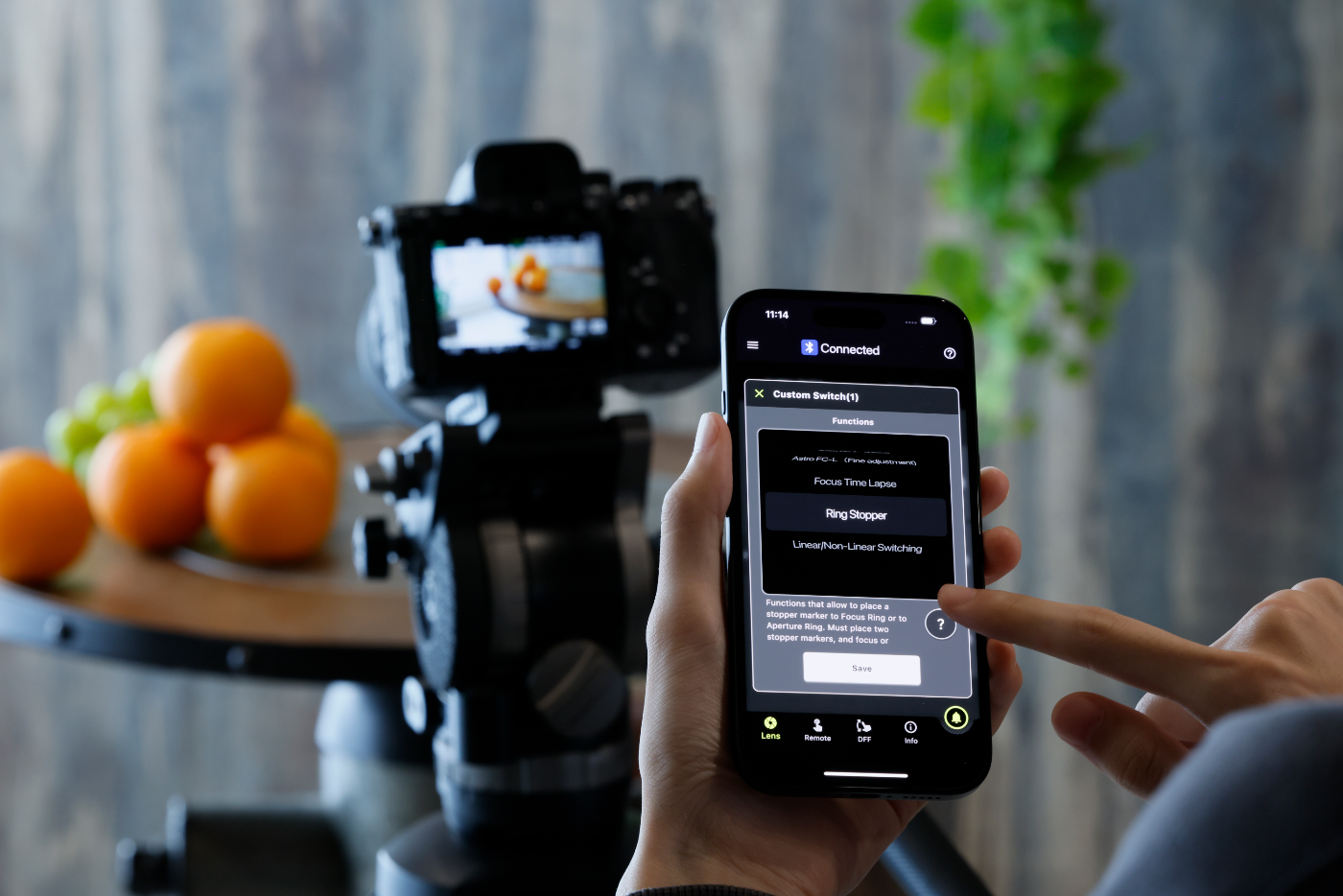 Hand houdt smartphone met Tamron Lens Utility-app voor draadloze lensbediening, met camera op statief op de achtergrond.
