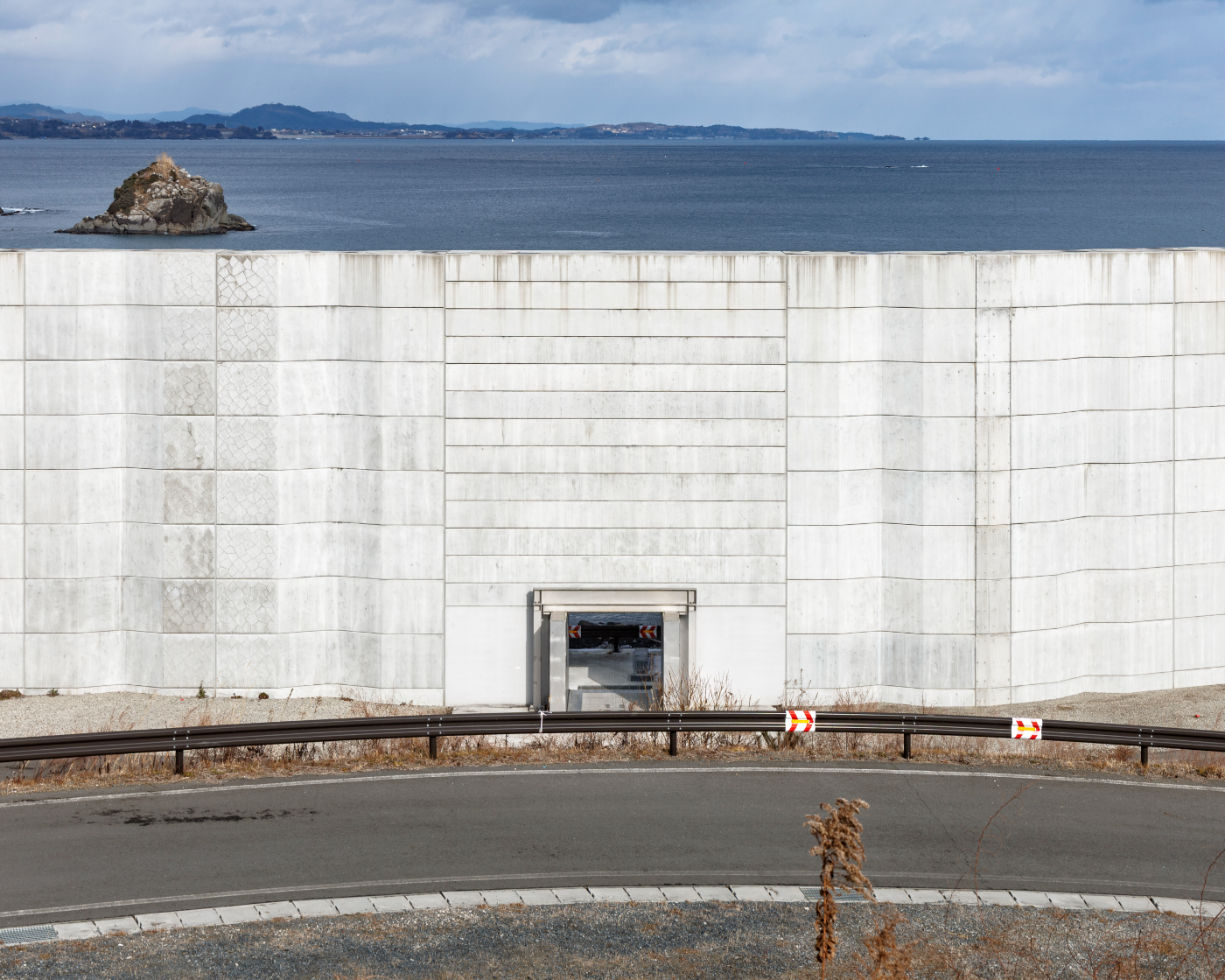 Strakke betonnen zeewering met kleine doorgang; voorgrond met weg en vangrail, op de achtergrond zee, rots en bewolkte lucht.