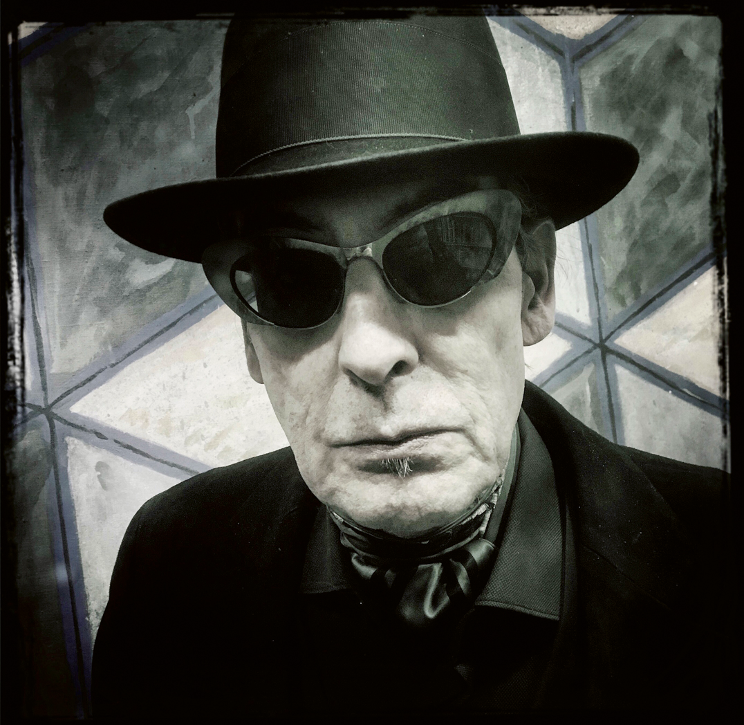 foto van de oudere jules deelder met hoed en zonnebril tegen een soort glaspatroon achtergrondin zwartwit