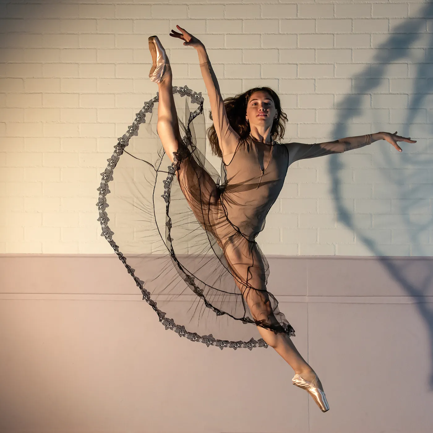 Balletdanseres Salome Leverashvili maakt een krachtige sprong tegen een lichte bakstenen wand, met een transparante rok die de beweging benadrukt en warm zijlicht dat haar houding accentueert. Foto: © Rob van Woerkom