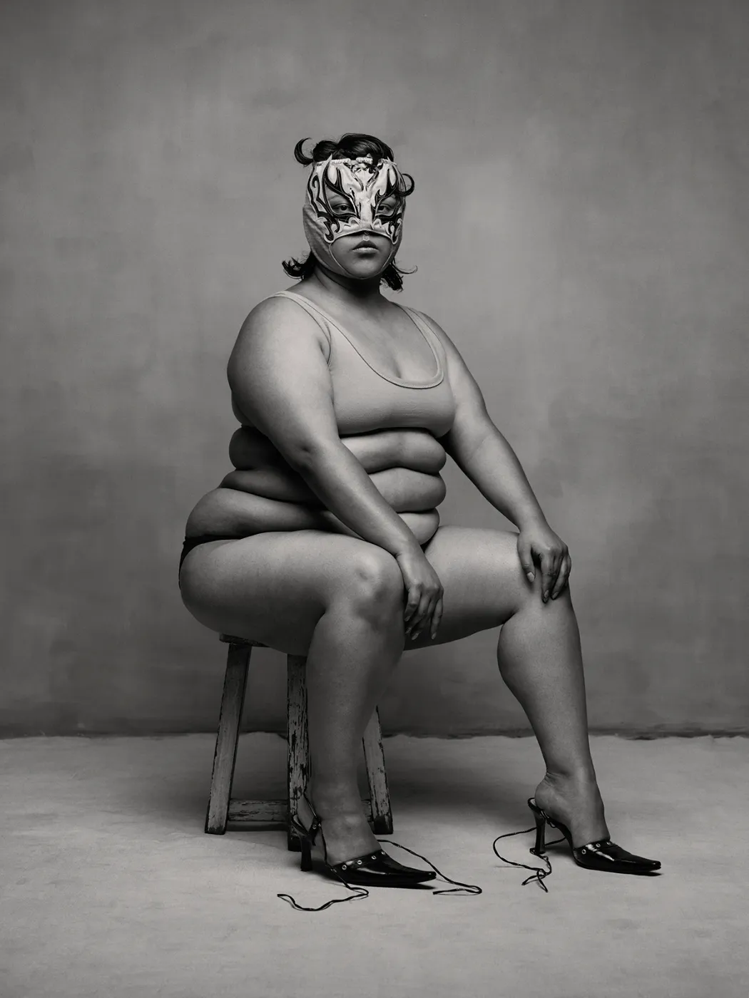 Zwart-wit portret van een vrouw met een luchador-masker, zittend op een houten kruk in minimalistische studio-opstelling, uit de serie Birds of Mexico City van Pieter Henket.