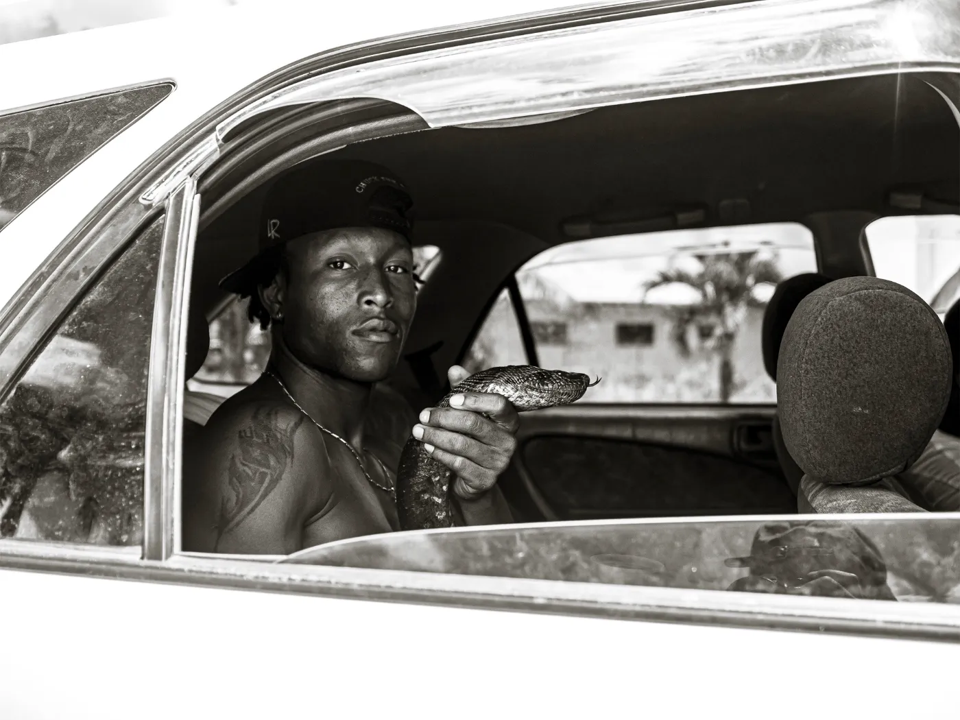 Zwart-wit documentair portret van een jonge man in een auto die een slang vasthoudt, gefotografeerd in rauwe straatfotografie-stijl door Stacci Samidin.