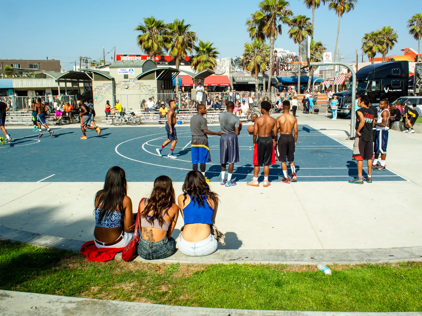 Straatbasketbalveld in Venice Beach met spelende basketballers, toeschouwers en palmbomen onder een zonnige Californische lucht.
