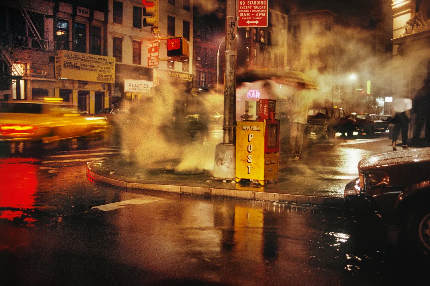 Straathoek in Manhattan bij avond met opstijgende stoom, nat asfalt, gele brievenbus en lichtsporen van passerend verkeer, gefotografeerd door Harry Gruyaert in 1985.