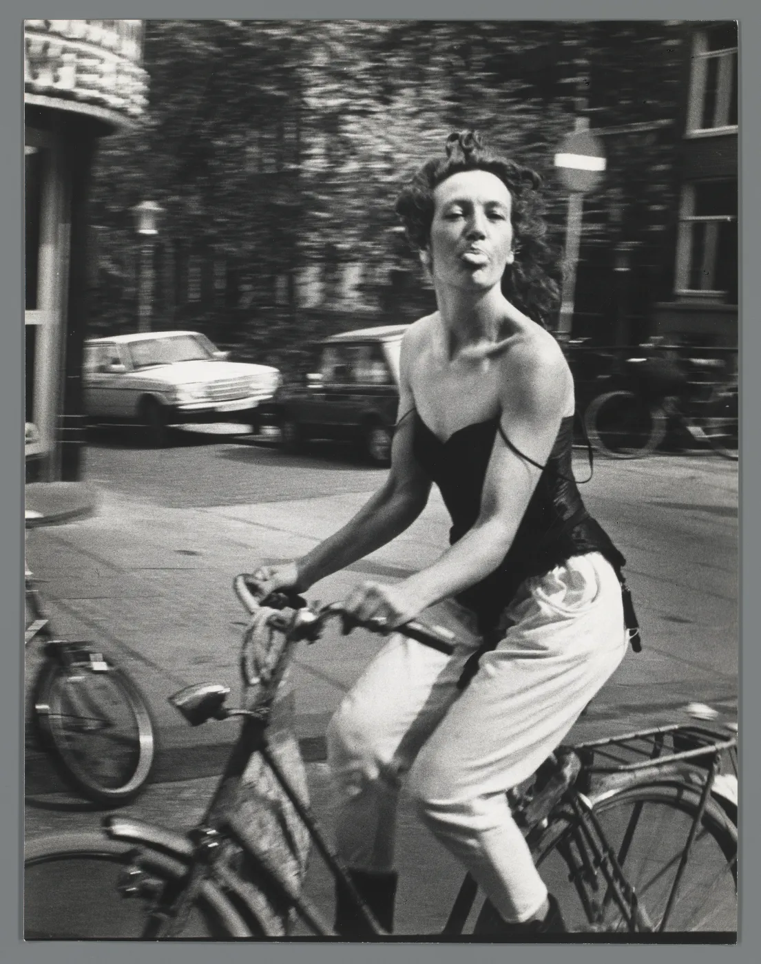 Ed van der Elsken, Woman on a Bicycle, 1983, Amsterdam. Zwart-wit straatfoto van een fietsende vrouw die speels haar tong uitsteekt naar de camera.