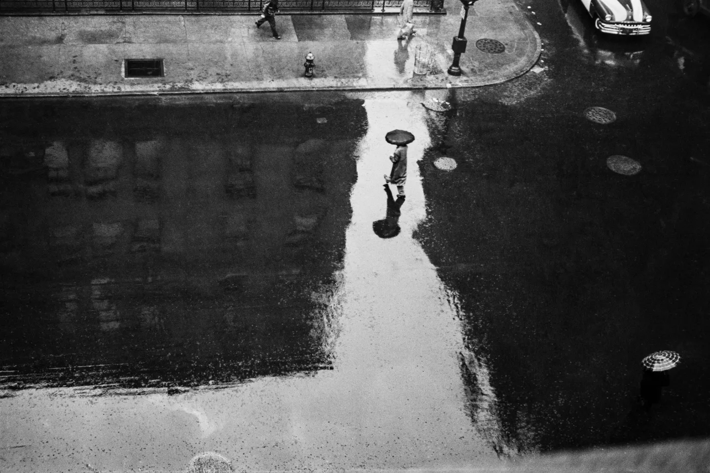 Zwart-witfoto van een natgeregende straat op Lexington Avenue, van bovenaf gefotografeerd, met een eenzame voetganger onder een paraplu weerspiegeld in het water.