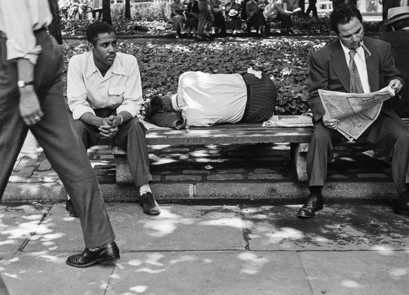 Zwart-witfoto van drie mannen op een bankje in Bryant Park: één man zit voor zich uit te kijken, een tweede ligt te slapen en een derde leest een krant met een sigaret in zijn mond.