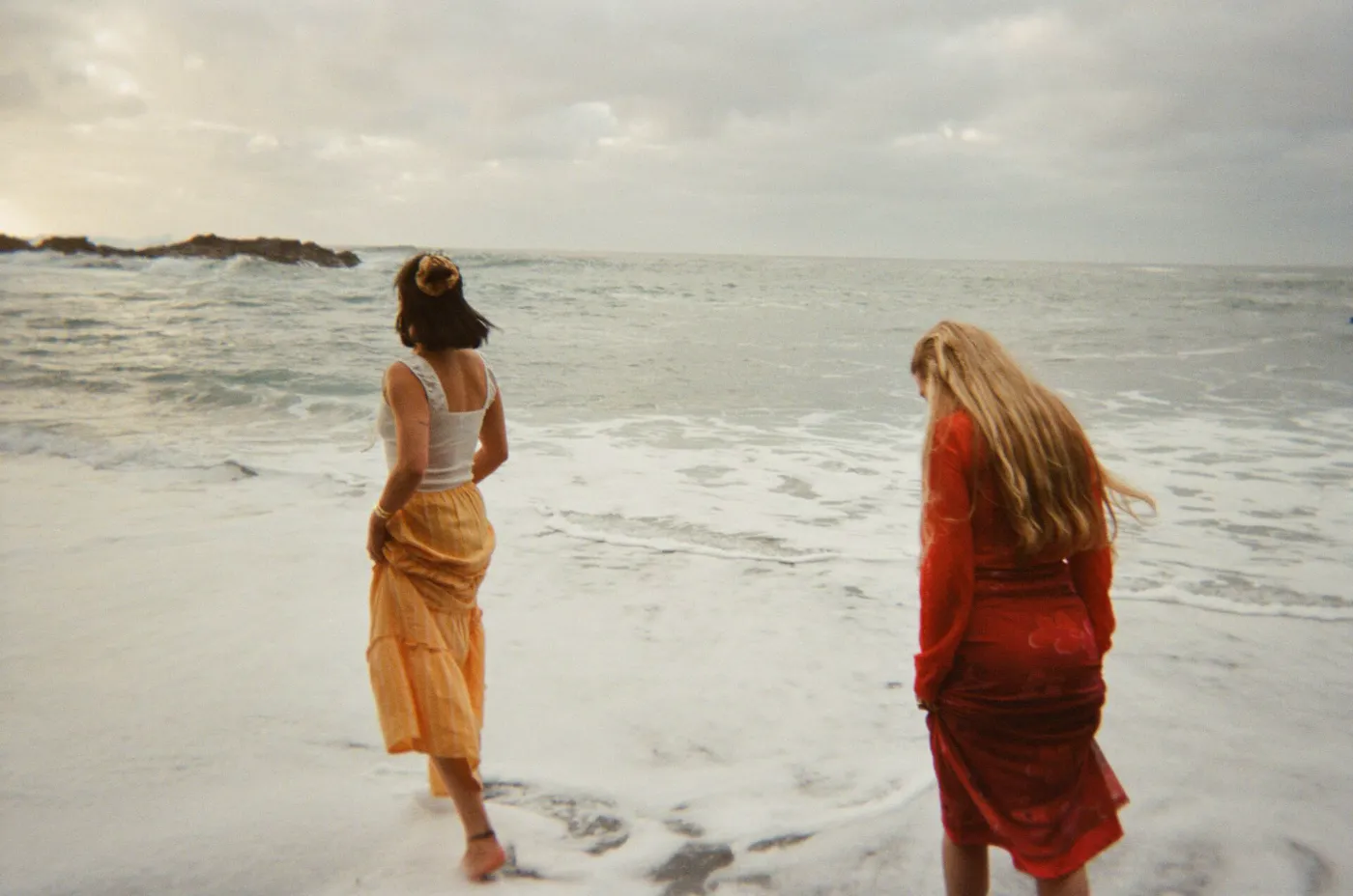 Twee vrouwen staan met hun voeten in de branding aan zee, gefotografeerd in zachte analoge sfeer